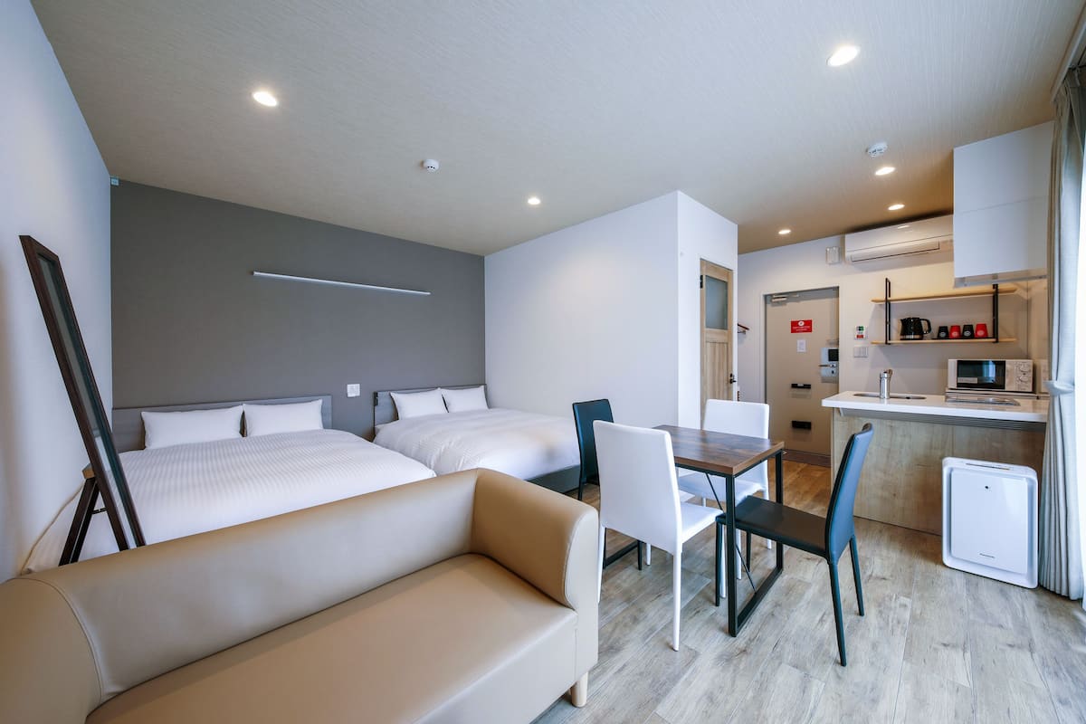 A modern hotel room with two beds, a sofa, a dining table with chairs, and a kitchenette.