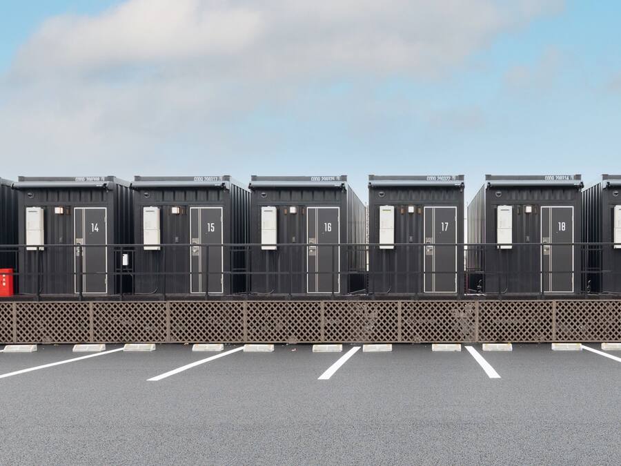 A row of six identical portable cabins with numbers 14 to 18, each with a door and a window, in a parking lot with a fence in the foreground.