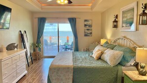 Un dormitorio con cama, cómoda, mesita de noche, lámpara y vista al mar a través de una puerta corrediza.