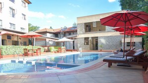 A swimming pool with sun loungers and umbrellas, surrounded by buildings with balconies and a clear sky. - Homes by Della (Nairobi)