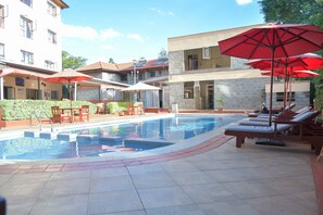A swimming pool with sun loungers and umbrellas, surrounded by buildings with balconies and a clear sky. - Homes by Della (Nairobi)