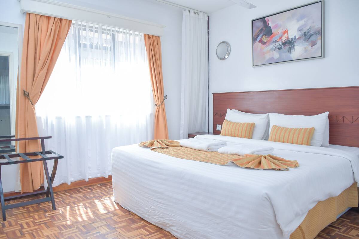 A hotel room with a bed, a wooden floor, a window with curtains, and a painting on the wall.