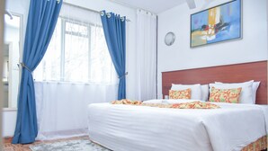 A neatly made bed with white linens and colorful pillows, a gray rug, and a window with blue curtains. - Homes by Della (Nairobi)