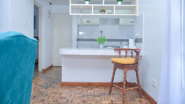 A compact kitchen with white cabinets, a wooden chair with a yellow cushion, and a herringbone-patterned floor. - Homes by Della (Nairobi)