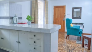 A kitchen with a white countertop, cabinets, and a potted plant. There is a wooden door and a blue chair in the background. - Homes by Della (Nairobi)