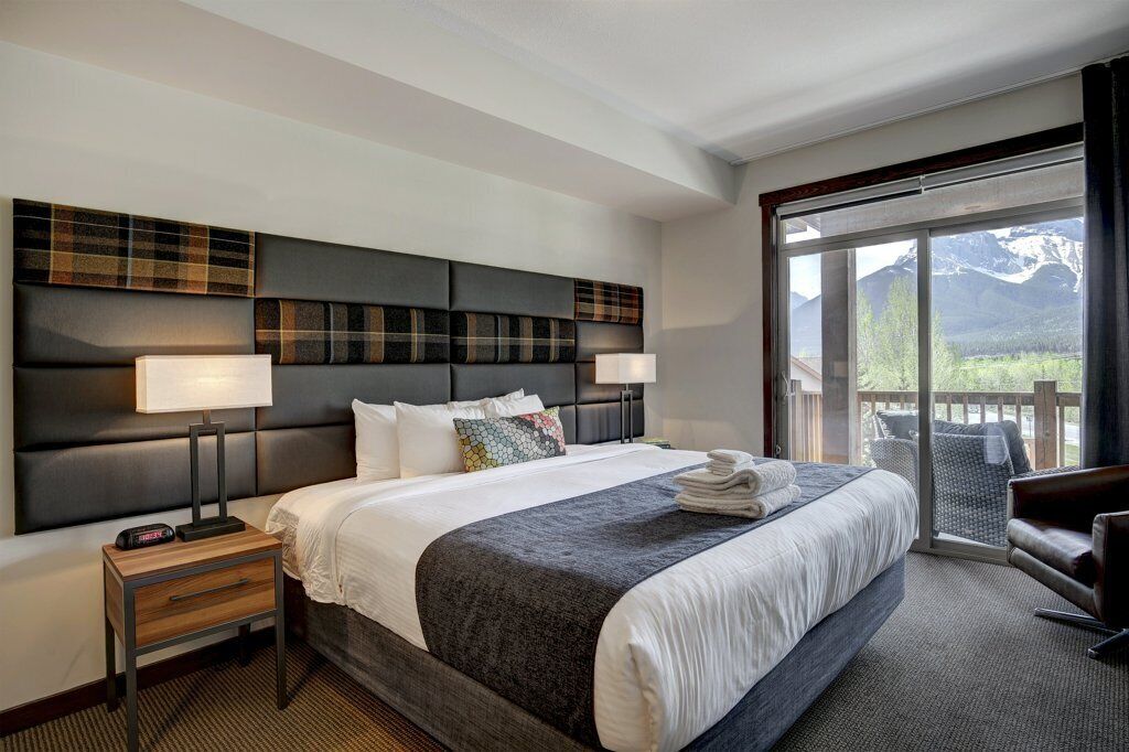 Habitación de hotel con una cama grande, una mesita de noche, una lámpara y vistas a las montañas a través de una puerta corrediza de vidrio.