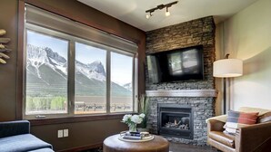 A living room with a stone fireplace, a flat-screen TV mounted on the wall, and a large window offering a mountain view. - Stoneridge 303 | Heated Outdoor Pool & Hot Tub (Canmore)