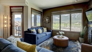 A modern living room with a blue sofa, a round coffee table, and a large window offering a mountain view. - Stoneridge 303 | Heated Outdoor Pool & Hot Tub (Canmore)