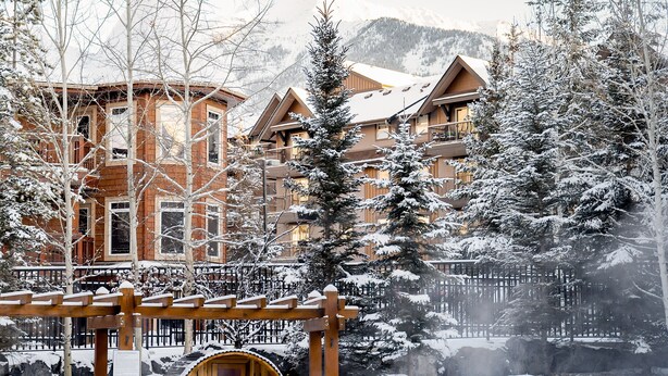 A snow-covered lodge with a hot tub, wooden deck, and a view of snow-covered mountains. - Stoneridge 303 | Heated Outdoor Pool & Hot Tub (Canmore)