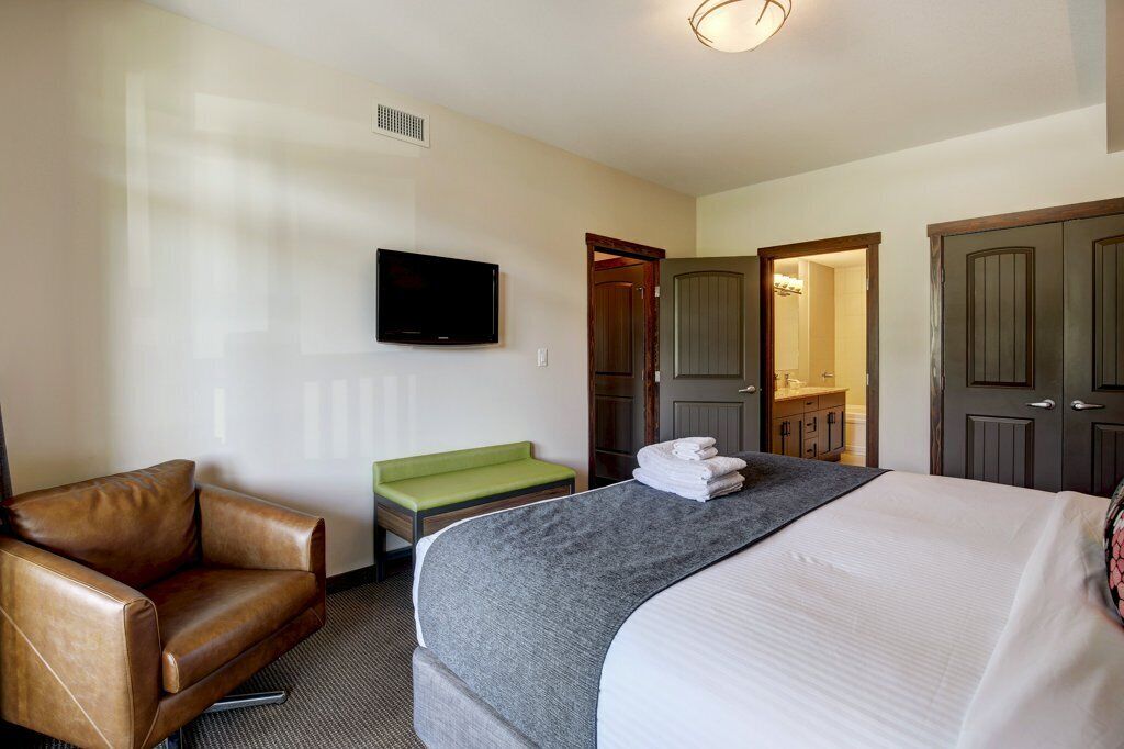 A hotel room with a bed, a leather armchair, a television, a green bench, and a bathroom visible through an open door.