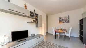 A modern living room with a flat-screen TV, a coffee table, and a dining area with a table and chairs. - City Apartment With Balcony and Parking (Lugano)