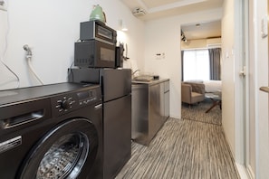 A compact hotel room with a washing machine, a compact kitchenette, a bed, and a small seating area.