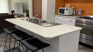 Moderni keittiö, jossa on valkoinen työtaso, ruostumattomasta teräksestä valmistettu pesuallas ja sisäänrakennettu mikroaaltouuni. Keittiössä on kolme mustaa baarituolia ja ruostumattomasta teräksestä valmistettu liesi.