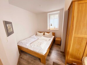 A bedroom with a wooden bed, a small bedside table, a window with blinds, and a wardrobe.