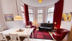 A modern living room with a dining area, featuring a white table, red chairs, a black sofa, and a red rug. - Vacation apartment Bucheneck 2 (Graal-Müritz)
