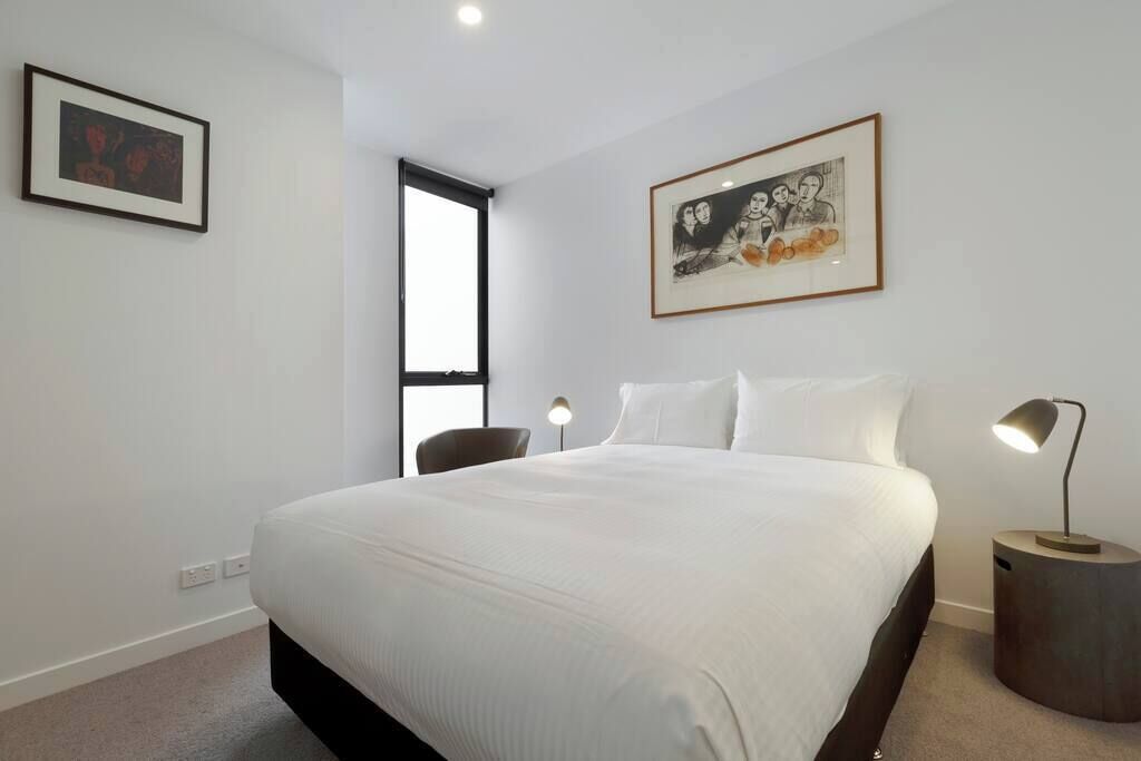 A bedroom with a bed, a chair, a bedside table with a lamp, and two framed artworks on the wall.
