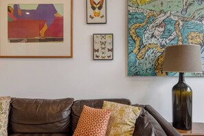 Un salon avec un canapé en cuir marron, des coussins à motifs, une œuvre d’art encadrée représentant des papillons, une lampe et une peinture abstraite aux couleurs vives accrochée au mur.