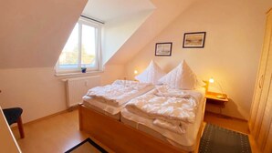 A bedroom with a sloped ceiling, a bed with white bedding, a bedside table with a lamp, and a window with a view.