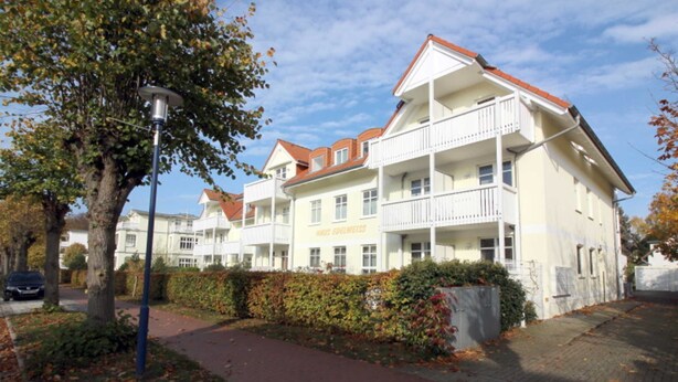 A multi-story hotel with a balcony on the second floor, a paved walkway, and a hedge in front.