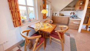A cozy dining area with a wooden table and chairs, a window with curtains, and a kitchen in the background.