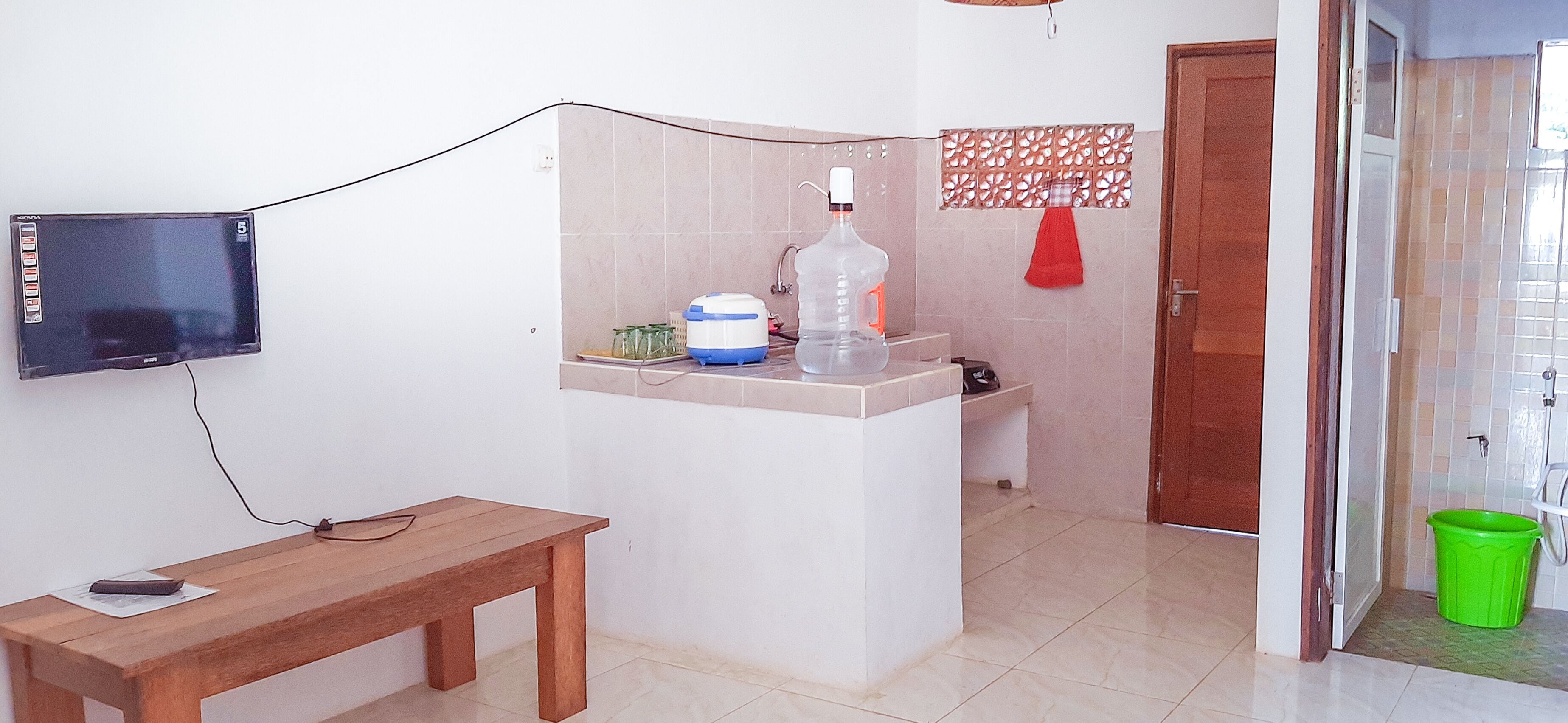 Une petite pièce propre, avec un banc en bois, un téléviseur fixé au mur, un lavabo et une poubelle verte.