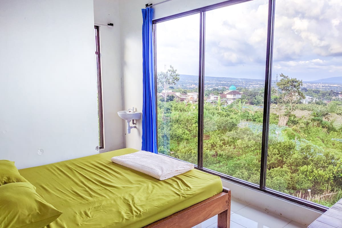 A bedroom with a large window, a bed with green bedding, a white sink, and a view of a green landscape.