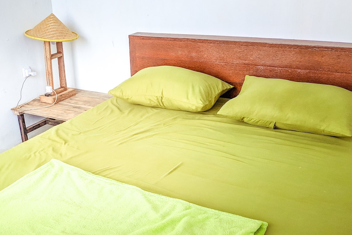A bed with green bedding and pillows, a wooden headboard, and a small bedside table with a lamp.
