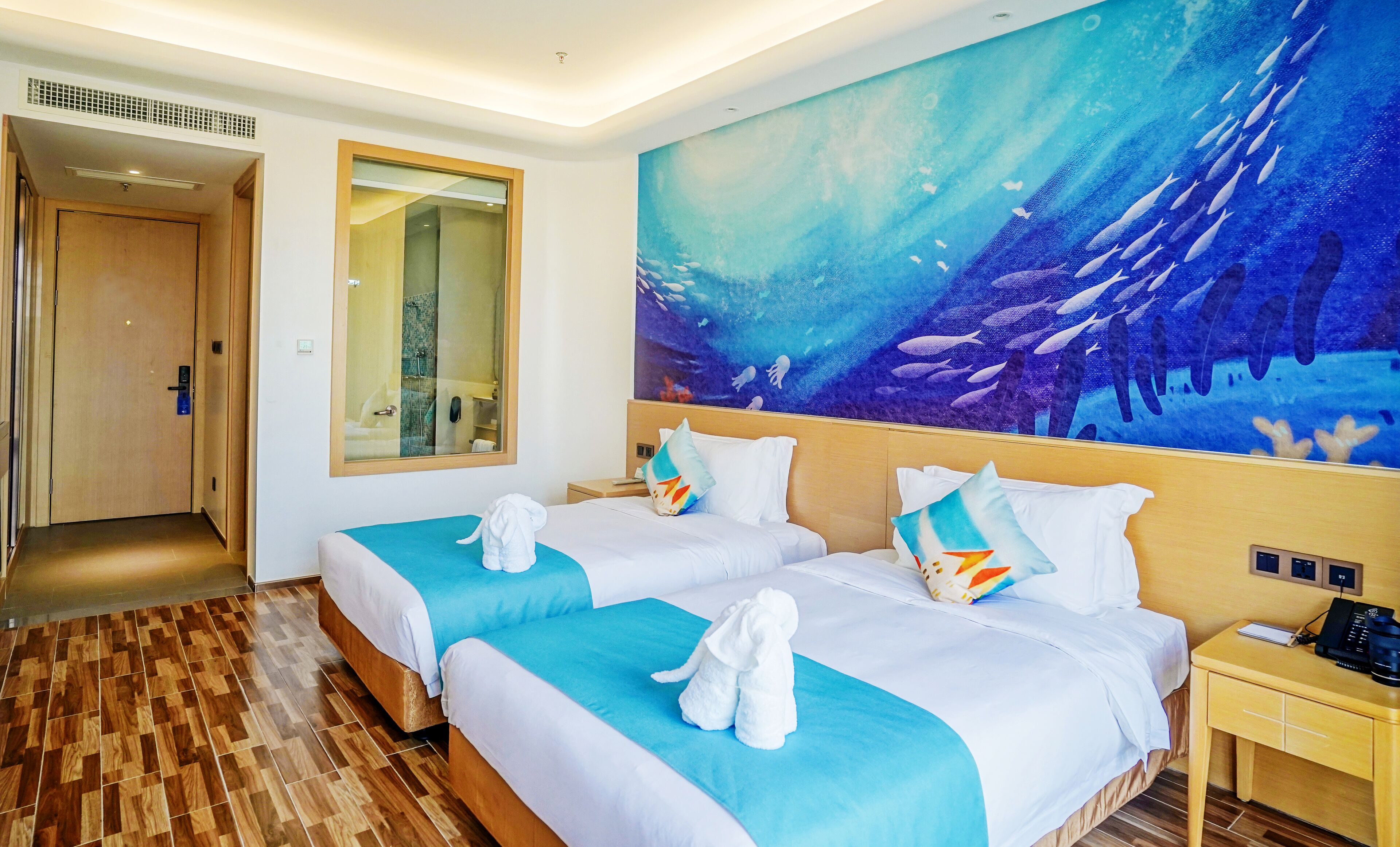 A hotel room with two beds, a large ocean-themed mural, a wooden floor, and a bathroom visible through a glass door.