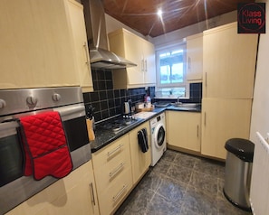 A compact kitchen with a stove, oven, and washing machine, a window with a view, and a trash bin. - Two Bedroom Apartment by Klass Living Serviced Accommodation Airdrie - Nicol Apartment With WiFi & Parking (Airdrie)