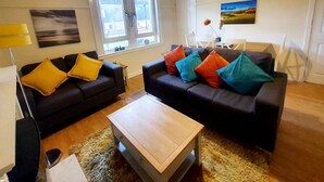 A living room with two sofas, a coffee table, and a lamp. There are windows, a painting, and a door. - Two Bedroom Apartment by Klass Living Serviced Accommodation Airdrie - Nicol Apartment With WiFi & Parking (Airdrie)