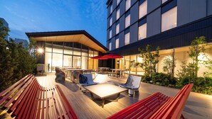 Un patio al aire libre con bancos y mesas de madera, rodeado de follaje y un edificio moderno con amplias ventanas.