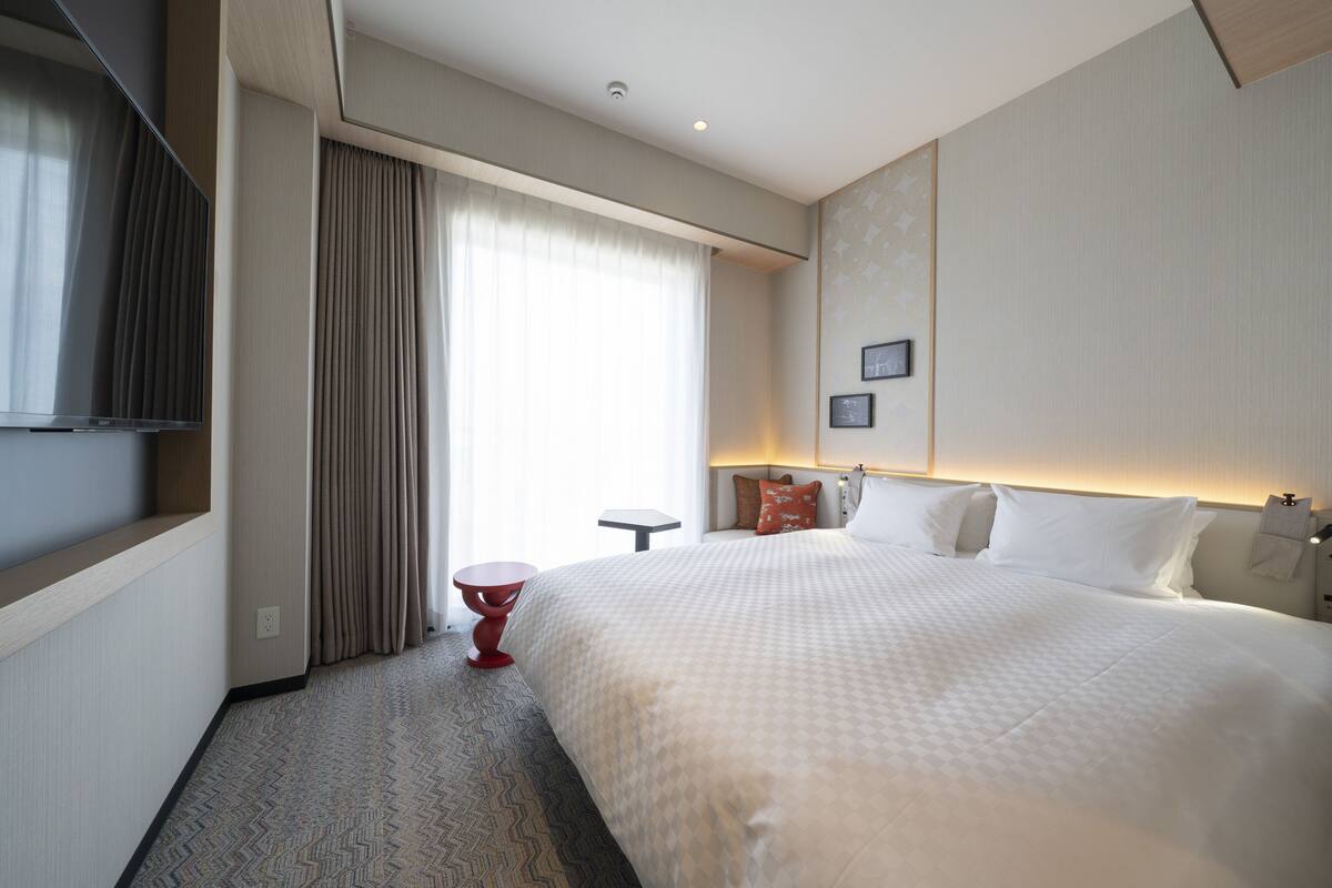 A hotel room with a large bed, a TV, a small table, and a window with curtains.