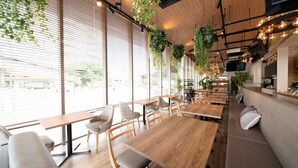 Un café moderno con mesas y sillas de madera, ventanas amplias con persianas, plantas colgantes y una zona de bar.