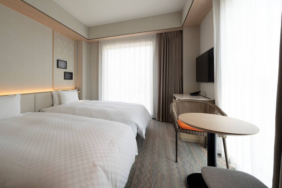 A hotel room with two beds, a desk, a chair, a television, and a large window with curtains.