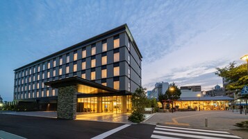 Un hotel moderno con fachada de vidrio, base de piedra y un paso de peatones enfrente.