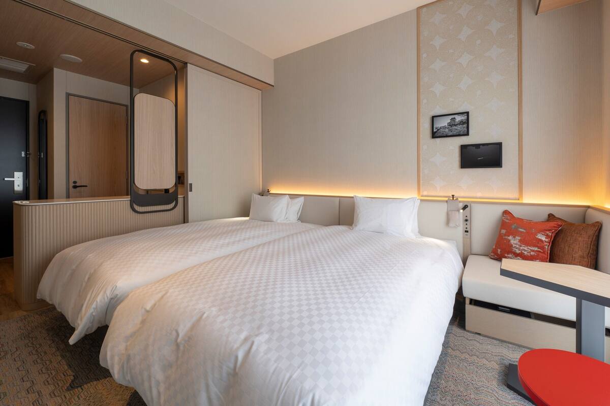 A modern hotel room with a large bed, a bedside table, a red stool, and a wall-mounted TV.
