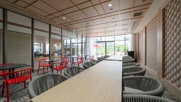 A modern conference room with a long table, red and grey chairs, and large windows offering a view of the outdoors.