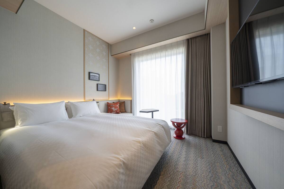 A hotel room with a large bed, a TV, a small table, and a window with curtains.