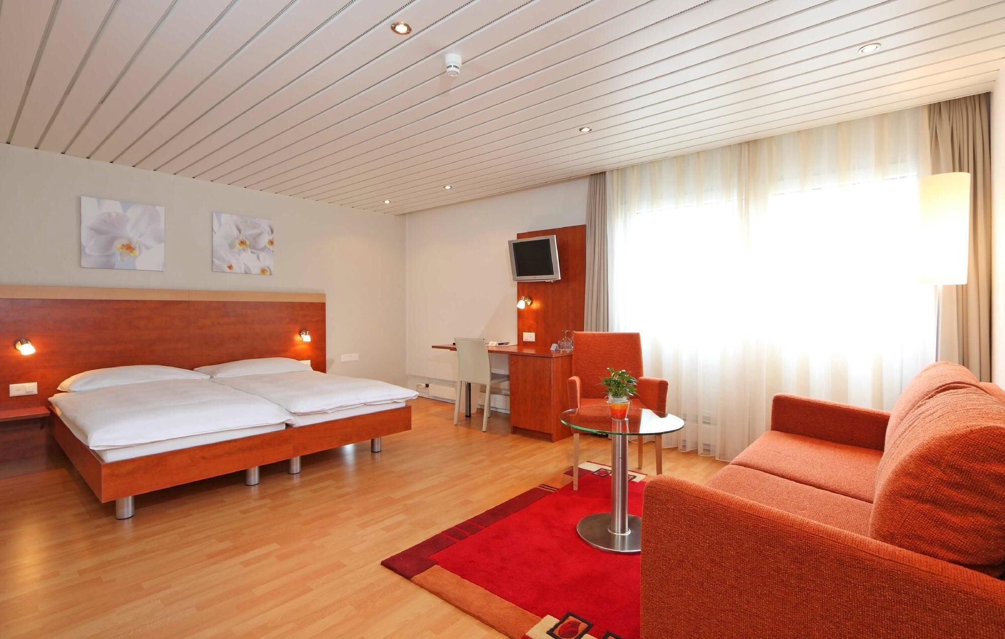 A hotel room with a wooden bed, a red sofa, a small table with a plant, a desk with a chair, and a TV mounted on the wall.