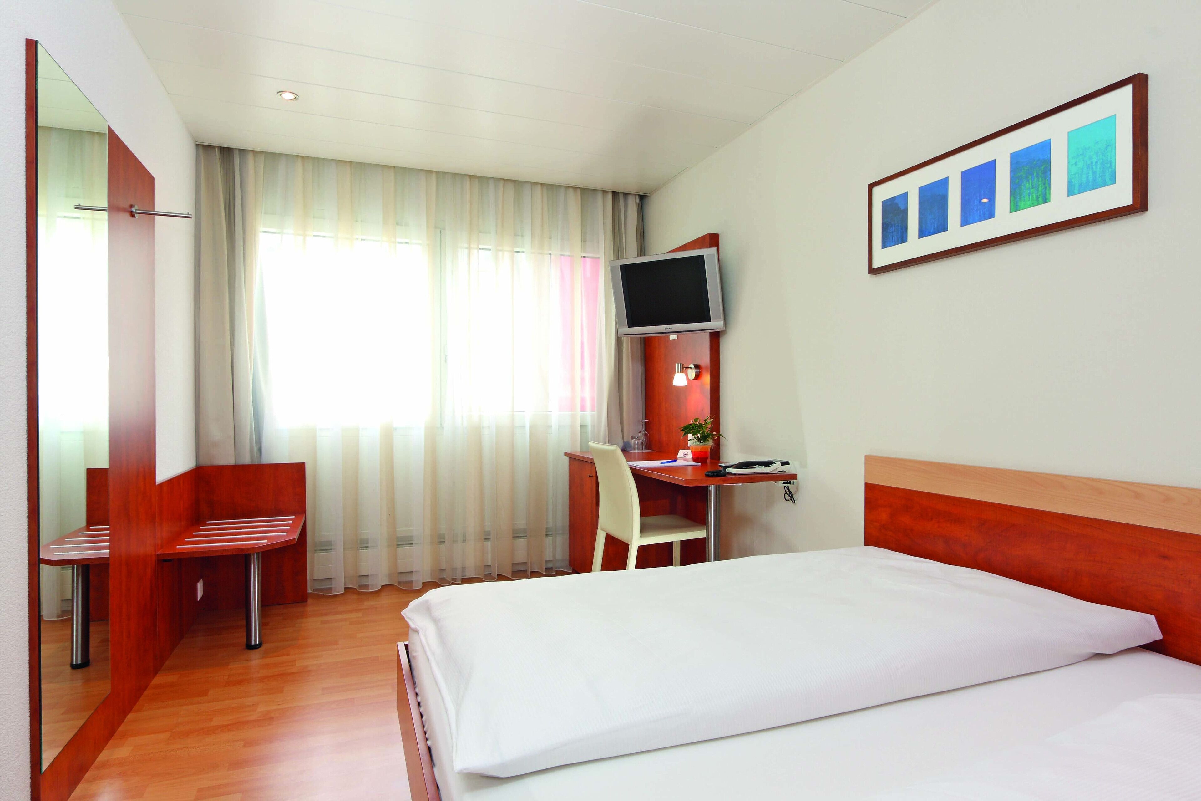 Une chambre d’hôtel comprenant un lit, un bureau avec une chaise, un téléviseur fixé au mur et une fenêtre avec des rideaux.