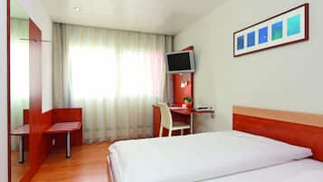Une chambre d’hôtel comprenant un lit, un bureau avec une chaise, un téléviseur fixé au mur et une fenêtre avec des rideaux.