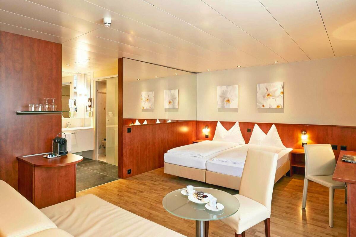 A modern hotel room with a bed, a seating area with a glass table, and a small kitchenette.