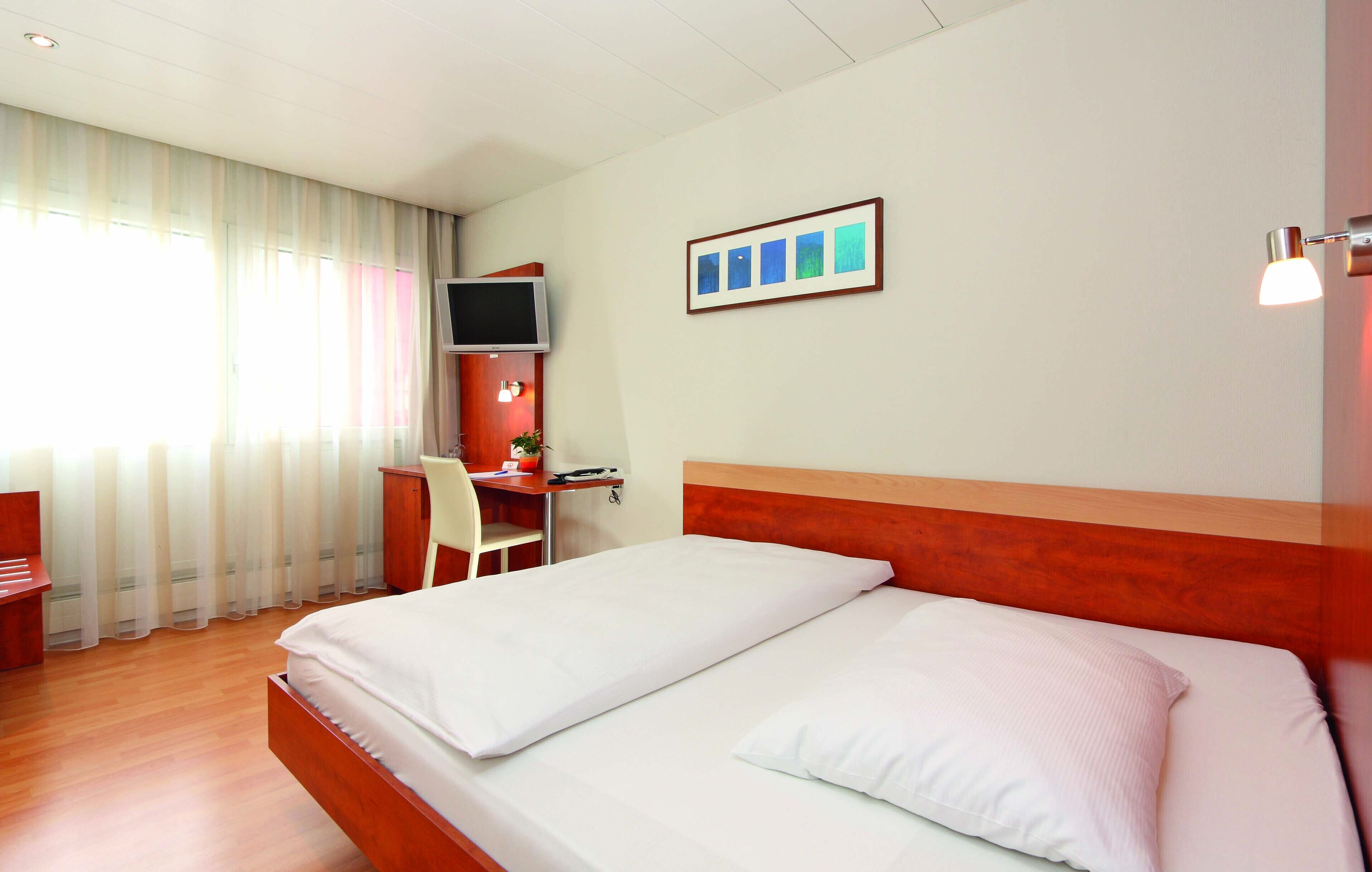 A hotel room with a bed, a desk, a chair, a TV, and a framed picture on the wall.