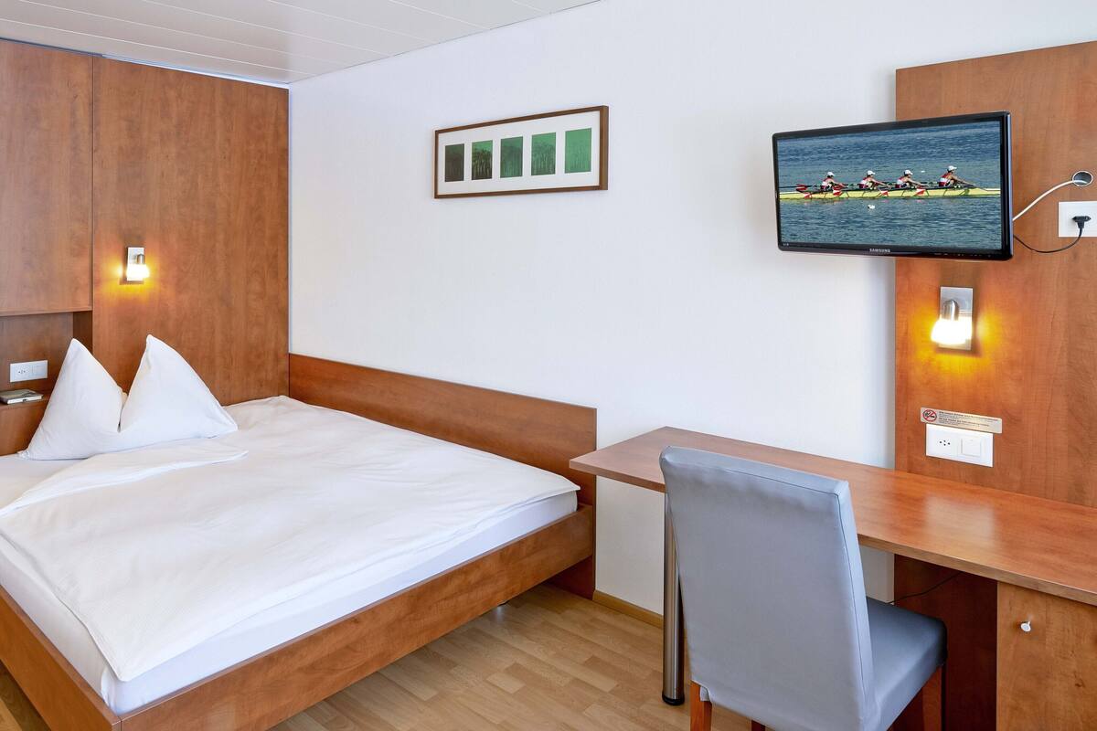 A hotel room with a wooden bed, a desk with a chair, a television mounted on the wall, and a framed picture above the bed.