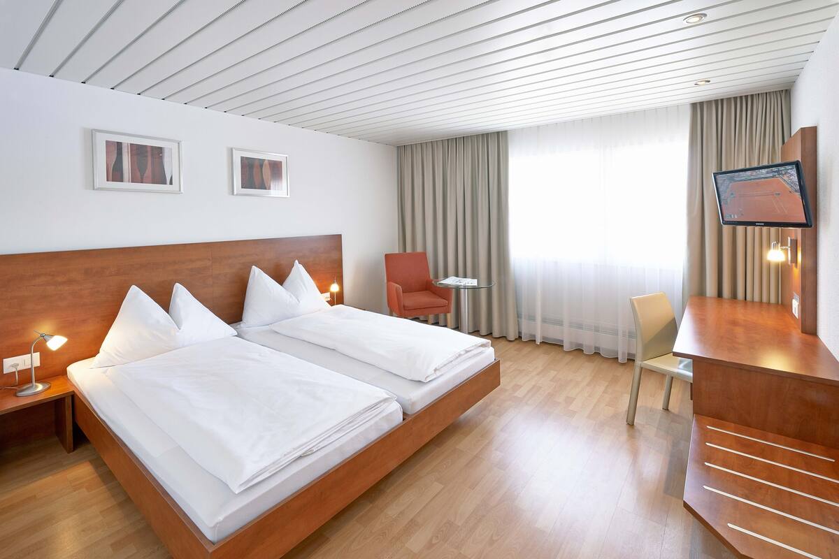 A hotel room with a wooden bed, a desk, a chair, a TV, and a window with curtains.
