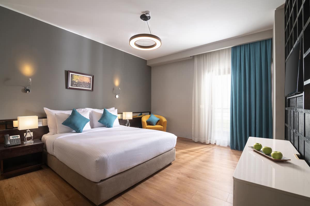 Moderni hotellihuone, jossa on suuri sänky, yöpöydällä lamput, työpöytä, jossa on omenoita, ja ikkunasta avautuva näkymä kaupunkiin.