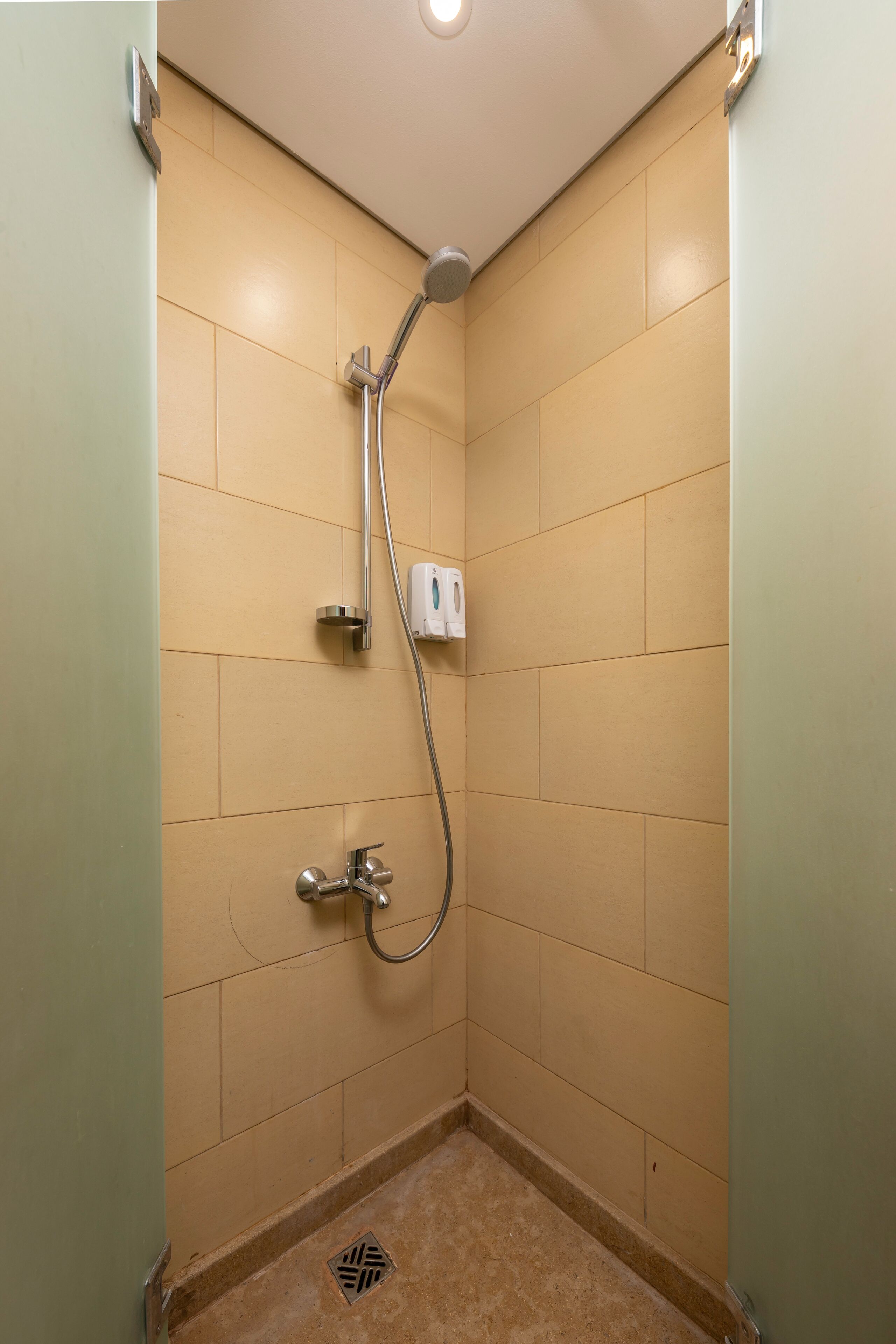 Een kleine, afgesloten douchecabine met beige tegels, een handdouche en een zeepdispenser aan de muur.