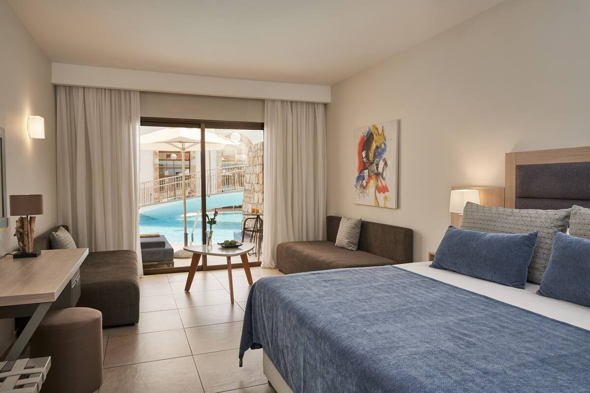 A hotel room with a bed, a desk, a chair, a small table with a plant, and a view of a pool and outdoor area.