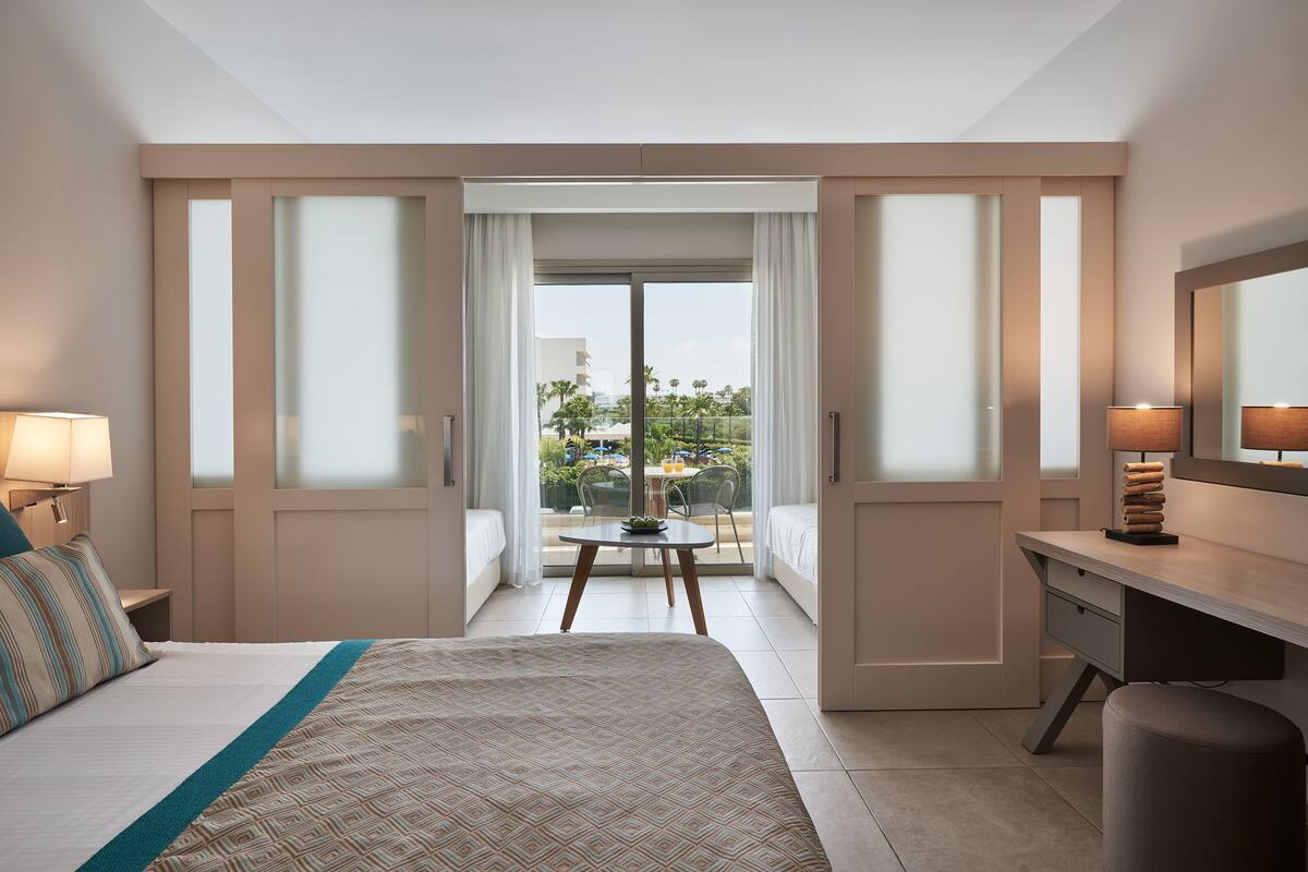 A hotel room with a bed, a desk, a chair, a table with a plant, and a balcony with a view of the city.