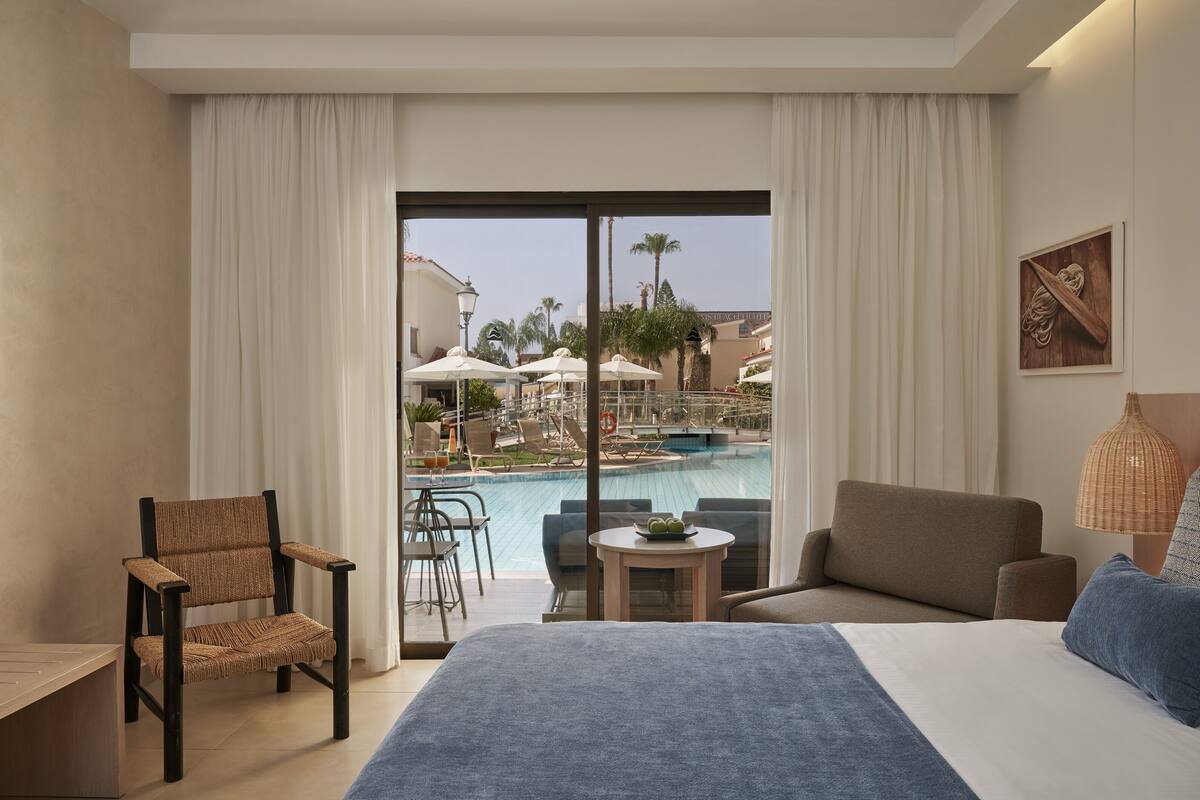 A hotel room with a bed, a chair, a small table, and a view of a pool and palm trees.